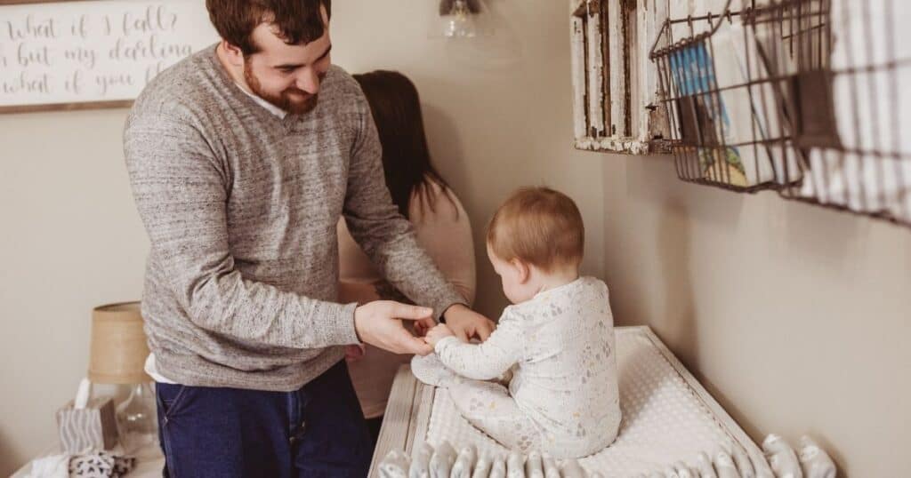 Dad holds babies hands while baby sits on changing table