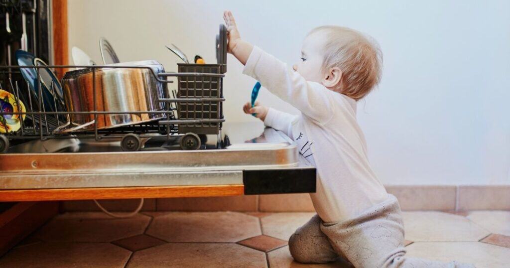 Baby sits on floor reaching past open dishwasher to grab cutlery out of rack