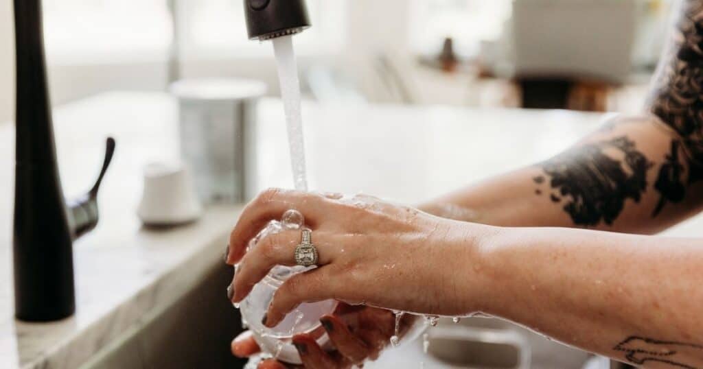Woman with tattooed arms washes bottled in soapy water with black faucet running