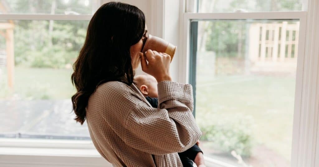 Mom in tan robe sips a cup of coffee while looking out the window and holding her baby