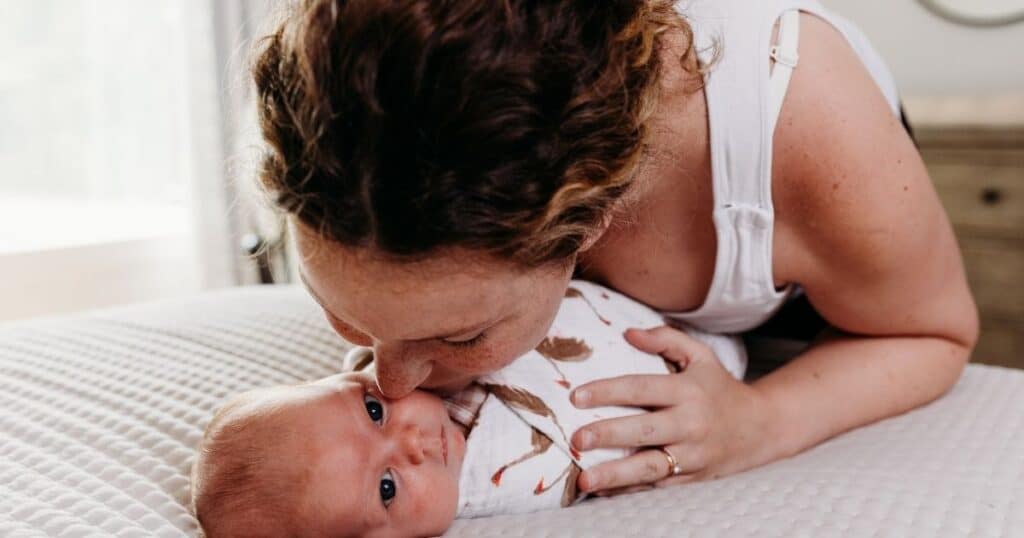 Mom leans over swaddled baby, kissing her on the cheek