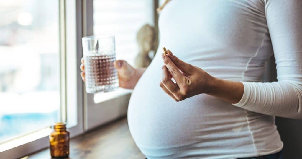 Pregnant woman stands at window with glass of water in one hand and a prenatal vitamin in the other