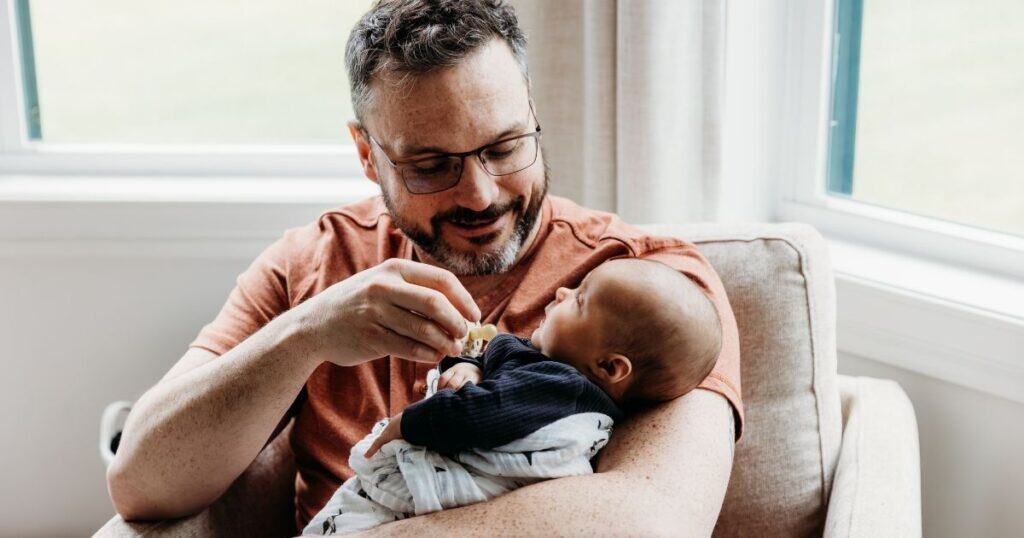 Dad sitns on chair in well lit room with his baby in his arms