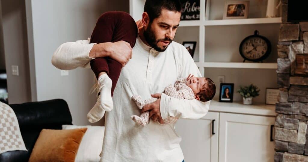 Father carries one child over shoulder while cradling newborn in other arm