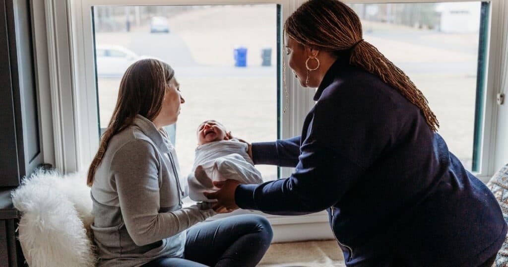 Postpartum Doula hands baby to mom sitting on bed