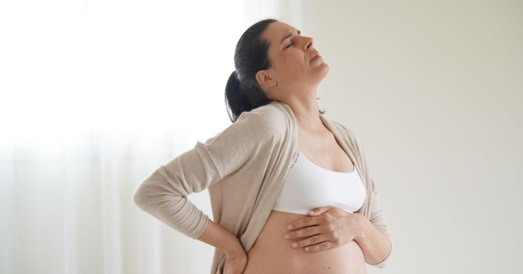 Pregnant woman in sports bra and cardigan sits on bed while holding her stoamch and back