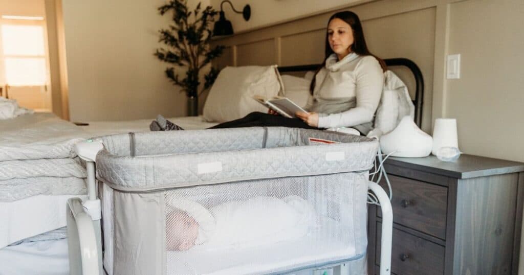 Mom relaxes in bed with book while baby sleeps in bassinet next to bed