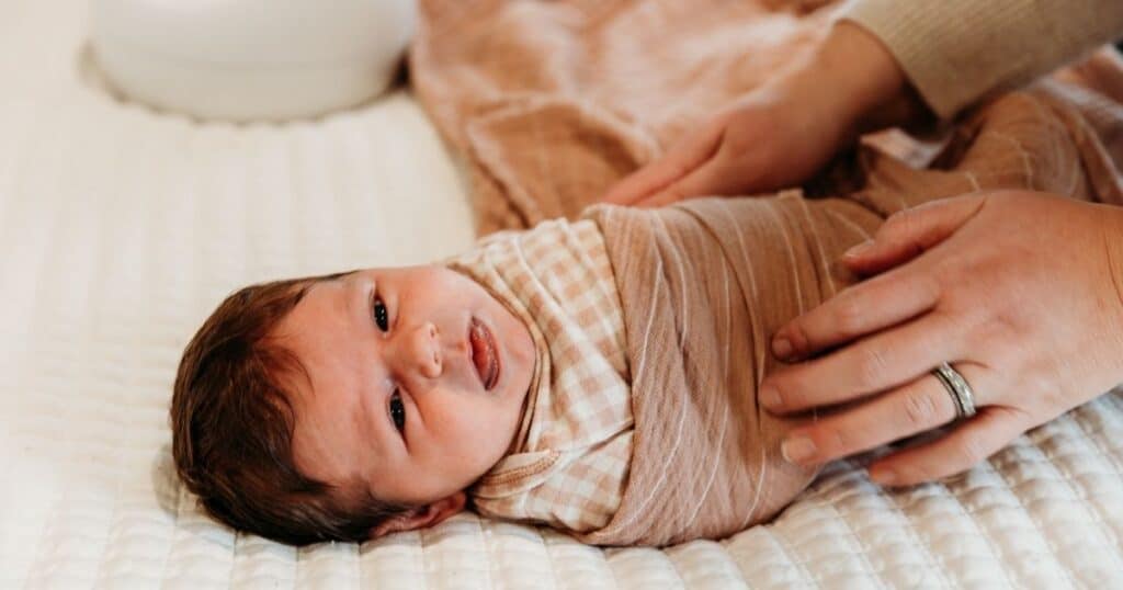 Baby lays on bed being swaddled while sticking thier tongue out