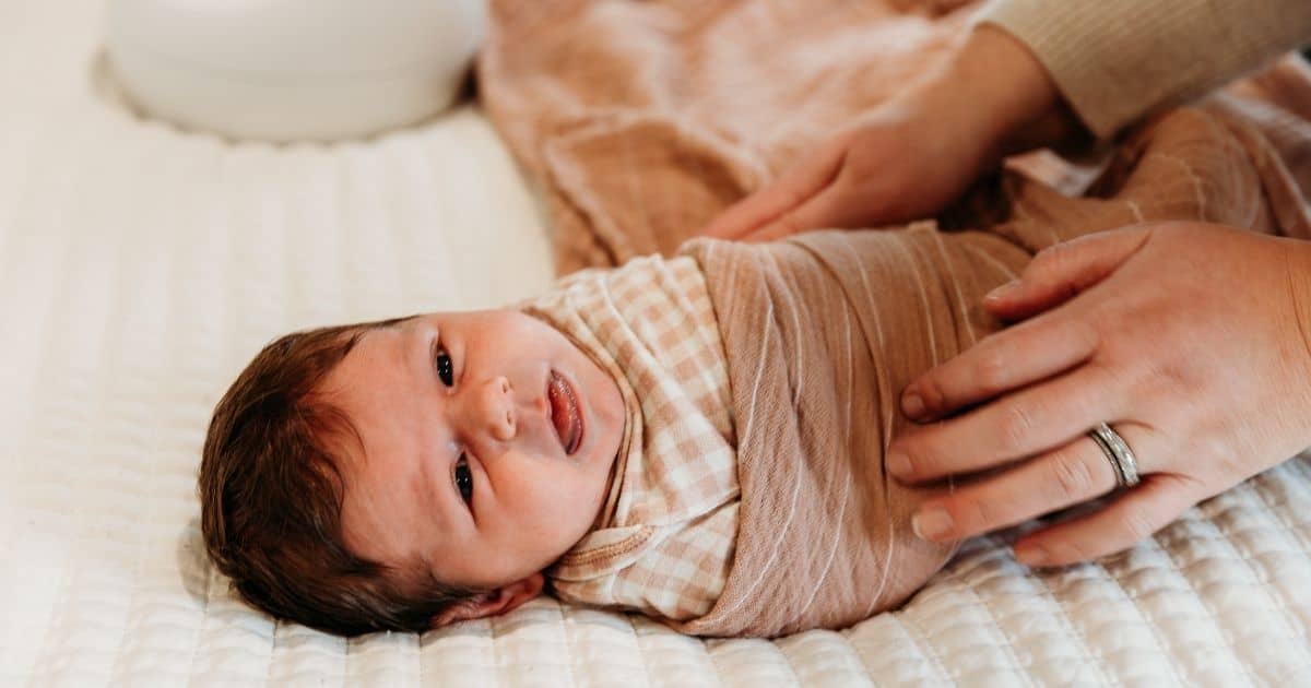 Baby lays on bed being swaddled while sticking thier tongue out