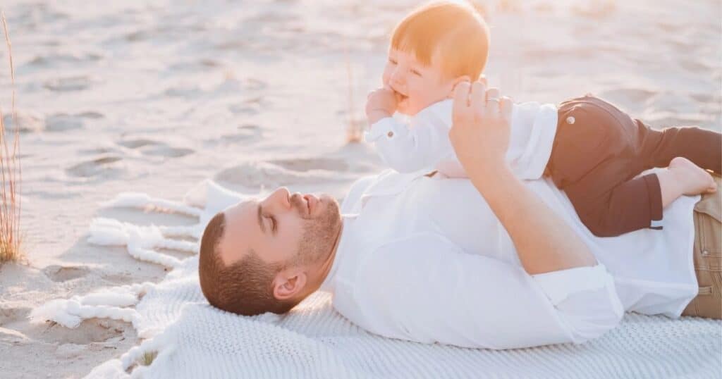 Day lays on blanket on the beach with baby on his chest
