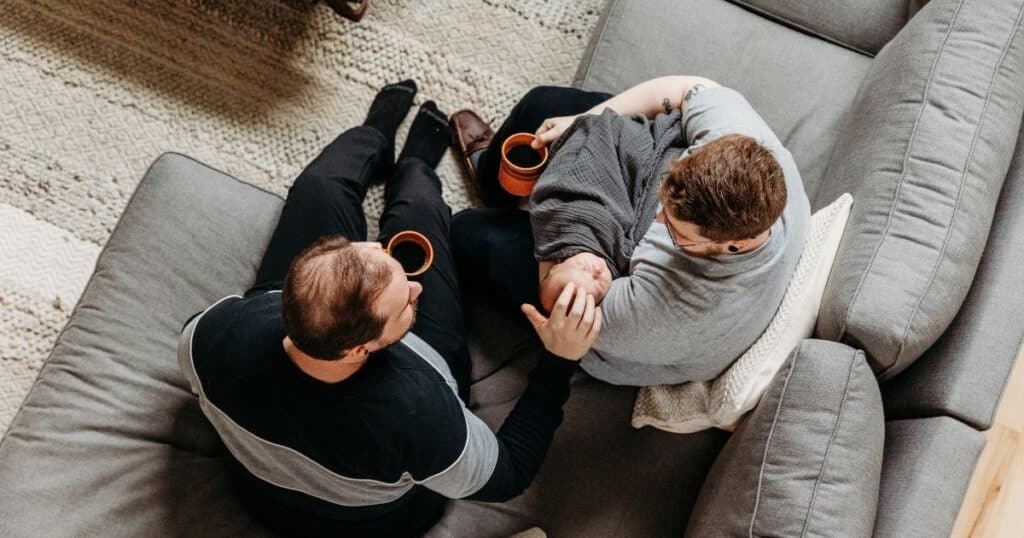 Photo looks down at two dads sittingon couch holding their baby and drinking coffee