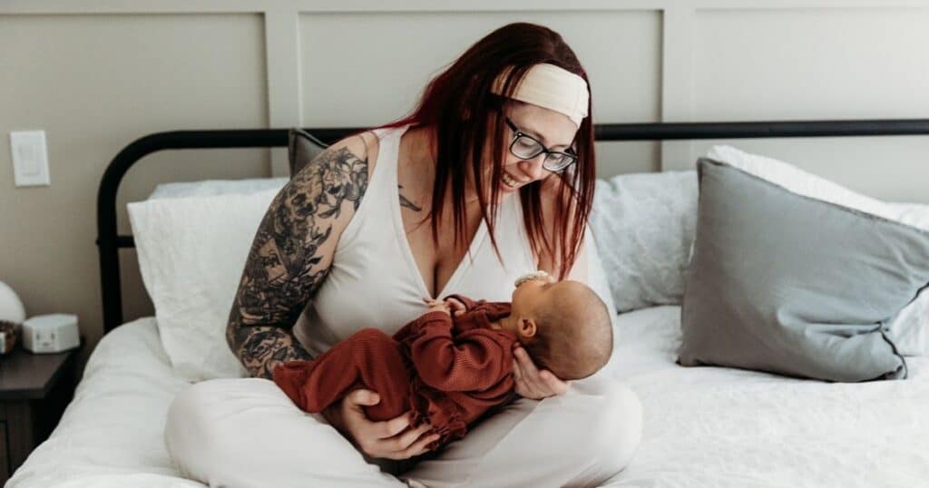 Mom with tattoos smiles down at her baby while sitting in bed