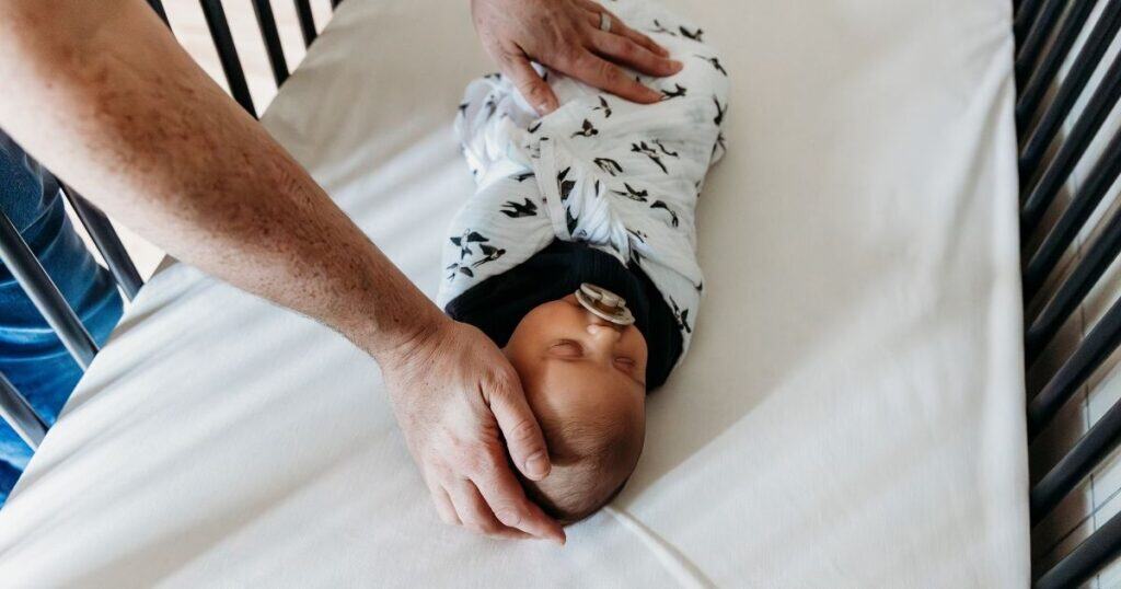 Baby in swaddle with pacifier sleepin in crib with parents hands on head and stomach