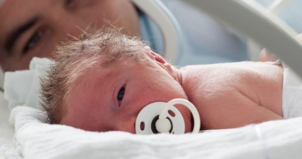 Preemie baby lays on stomach while using pacifier with mom in the background