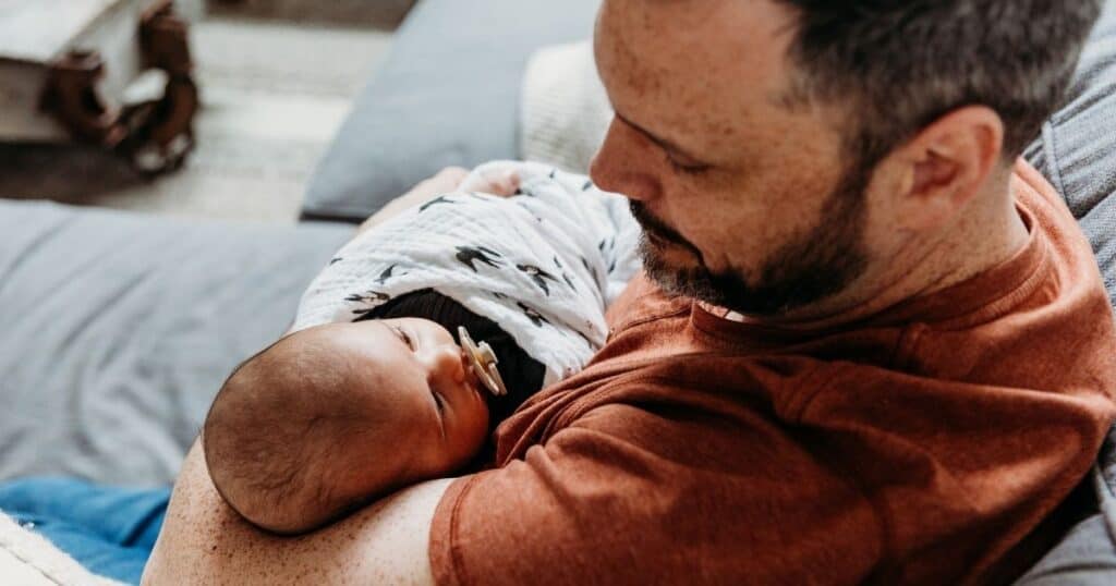 Freckled father holds swaddled baby as the look at eachother while sitting on the couch