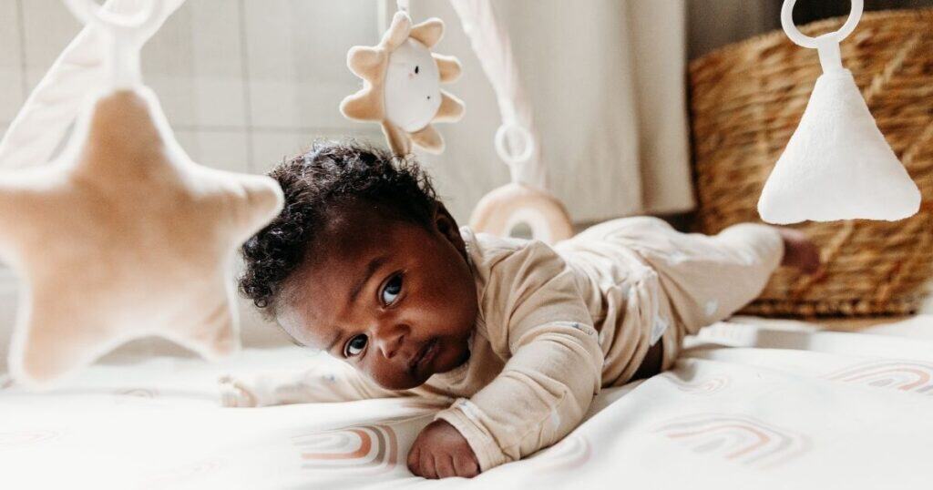 Baby in tan outfit does tummy time on a mat with sun and moon toys hanging from the top