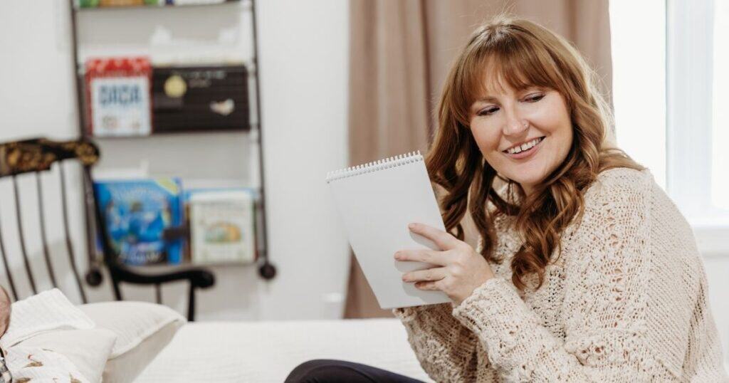 Woman sits on bed in nursery holding a notebook and pen