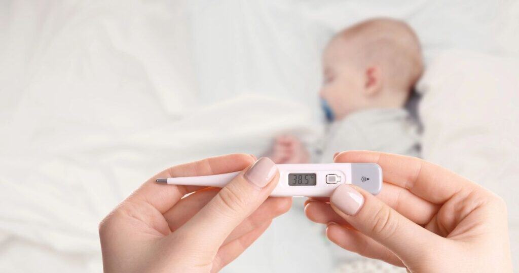 Mom holds thermometer while babt sleeps ion bed in the background