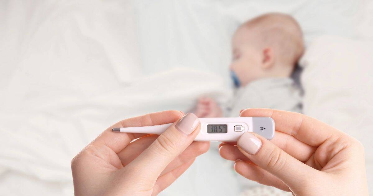 Mom holds thermometer while babt sleeps ion bed in the background