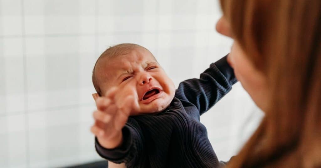Crying baby in dark grey onesie reaches out to Newborn Care Specialist