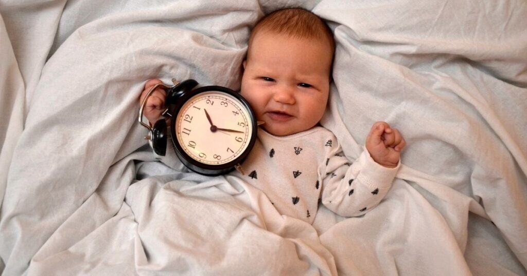 Baby lying in bed with tan blankets holding a black clock