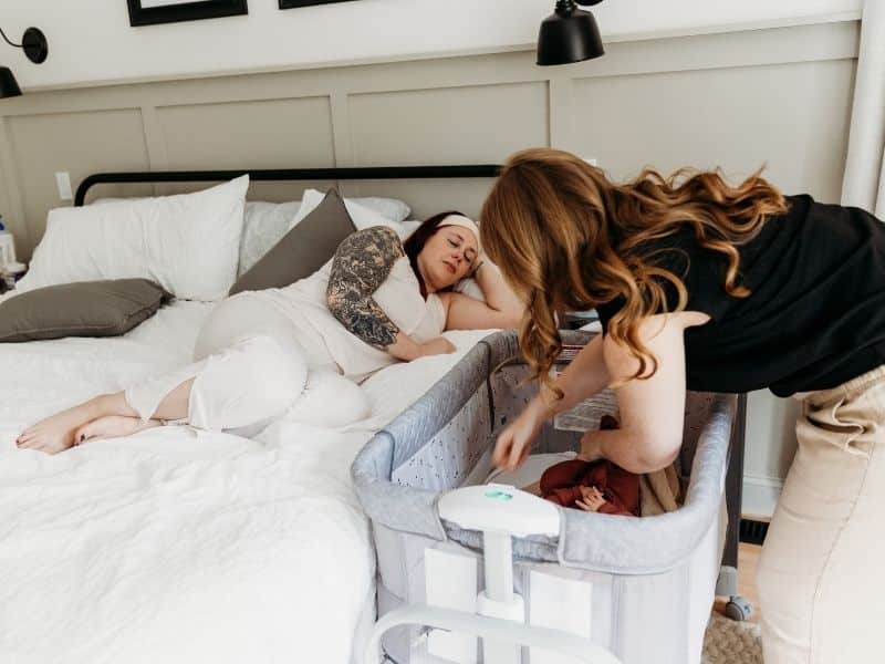 New mom resting in bed with doula by her side placing baby in the crib