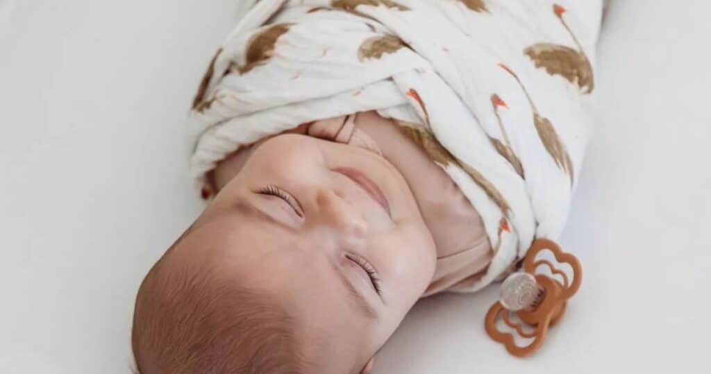 swaddled 2 month old baby with pacifier on flat mattress