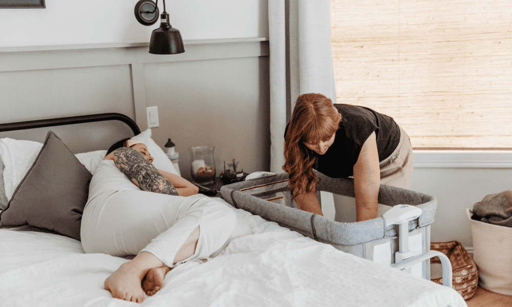 Newborn Care Specialist puts baby down to sleep with mom rests in bed next to bassinet