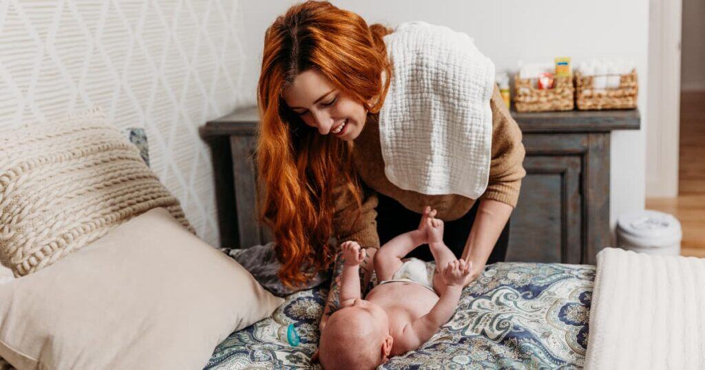 Newborn Care Specialist from Well Supported Family smiling at baby being dressed on bed.
