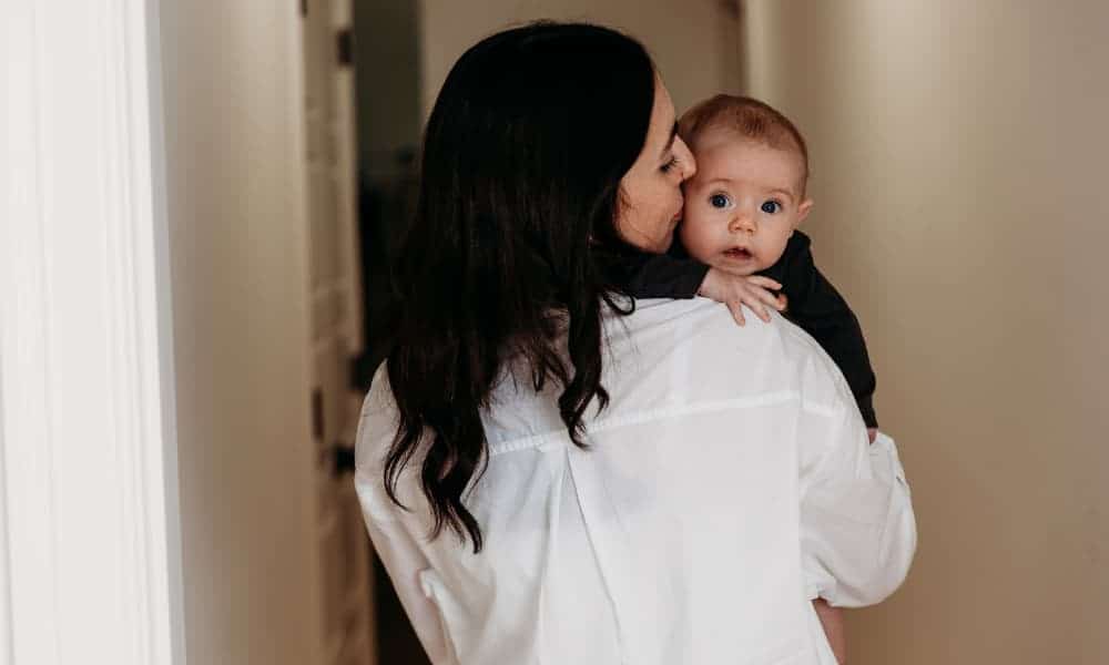 woman with dark hair wearing a white button-down shirt holds baby and kisses baby's head
