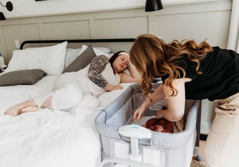 New mom resting in bed with doula by her side placing baby in the crib